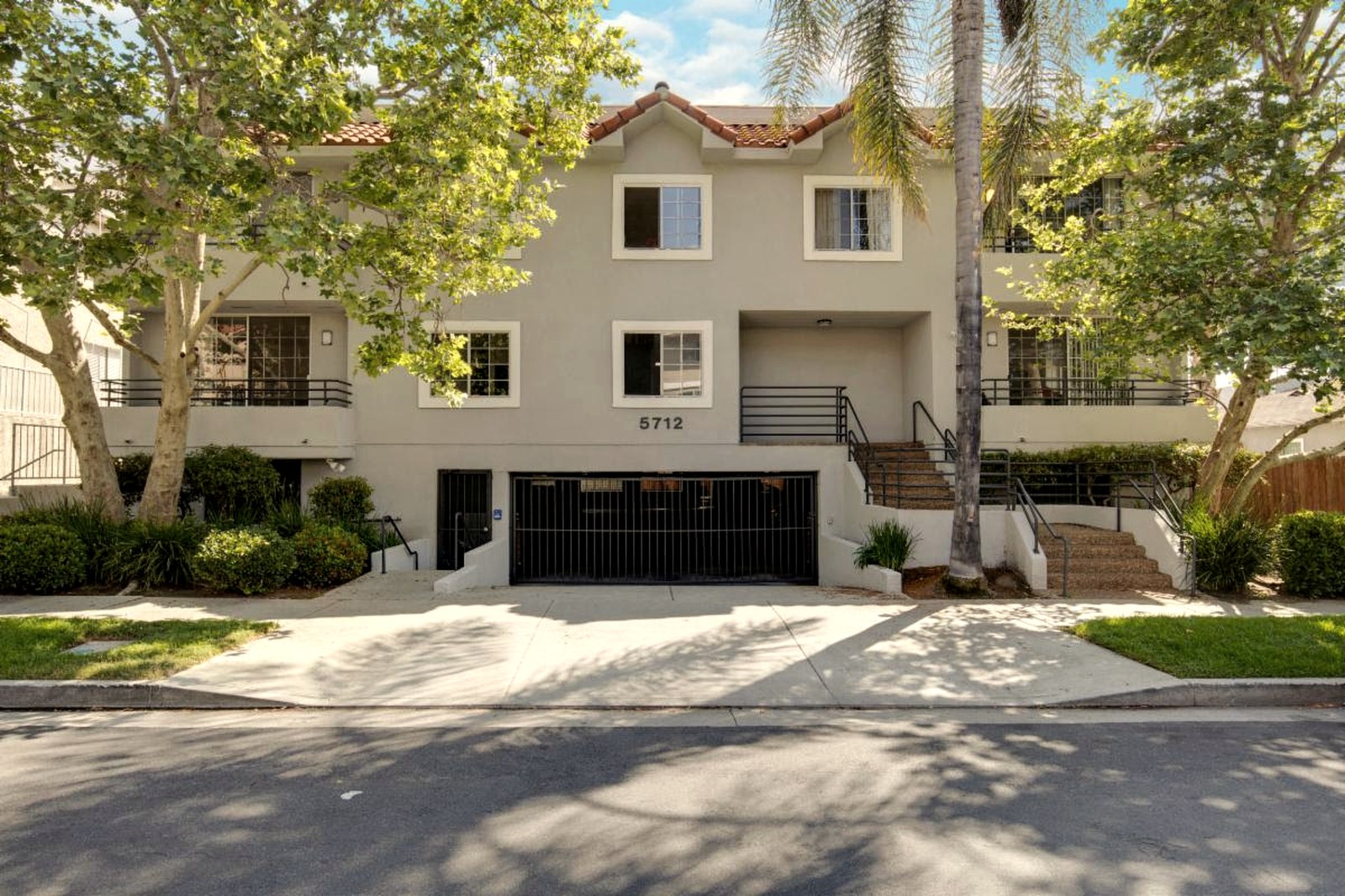 5712 Camellia Ave, a three-story stucco 14-unit apartment building in North Hollywood with arched parapets, gated tuck-under parking, mature palm trees, and a center address sign reading 5712.